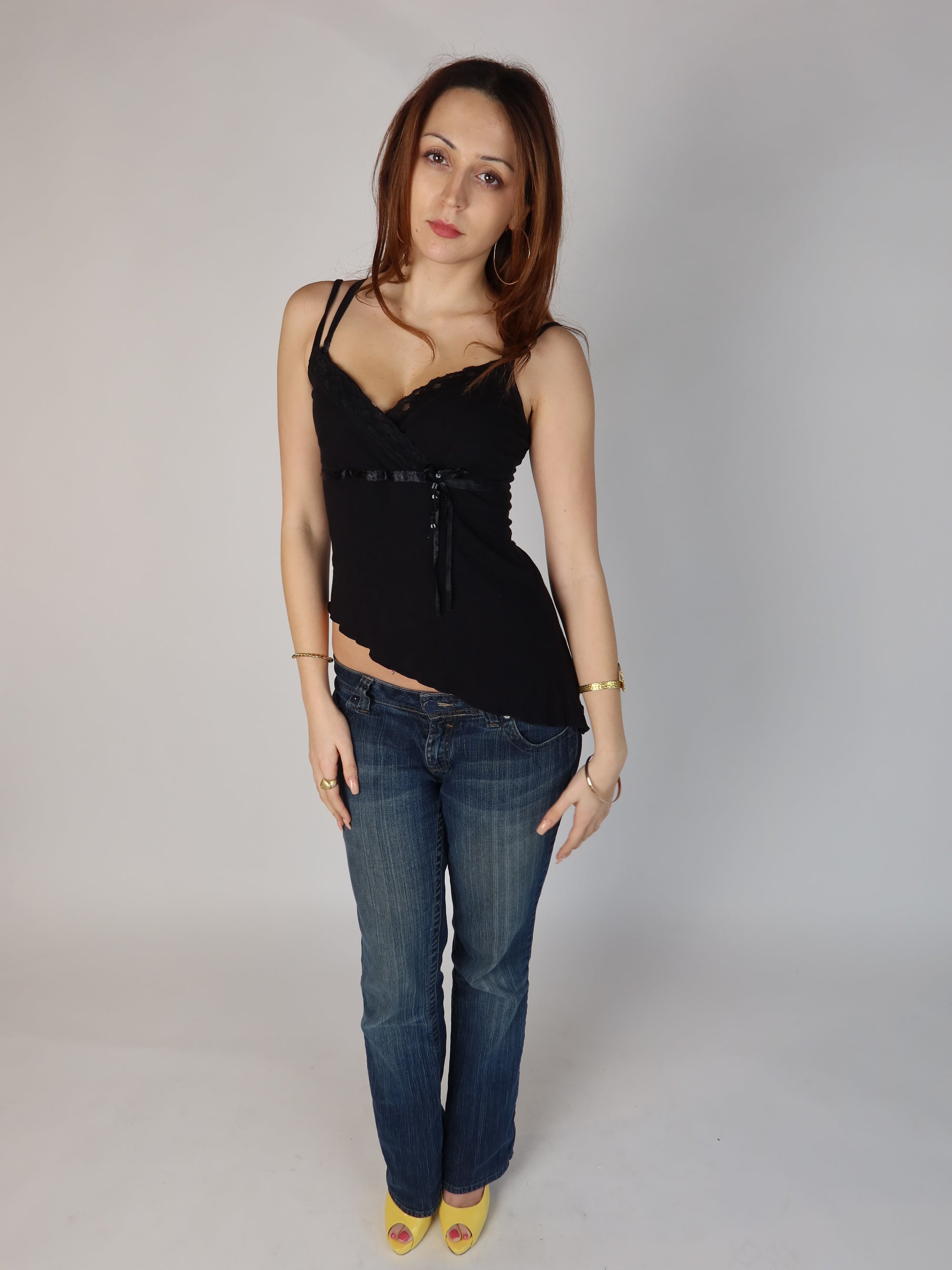 Authentic vintage Y2K black babydoll cami top with thin straps, lace trim cross over neckline, empire waist satin tie, and a slanted asymmetric hemline, shown from front and back views with low-rise jeans.