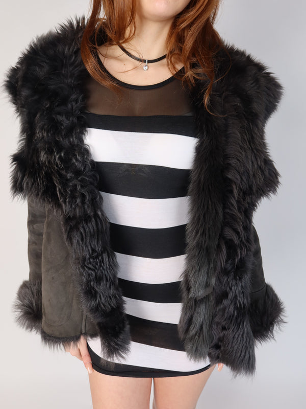 Authentic vintage Y2K premium black Italian shearling lambskin jacket featuring a completely fluffy fur-lined interior, an oversized shaggy fur collar, trim, and matching fur-cuffed sleeves, shown from front and back views with a striped mini dress.