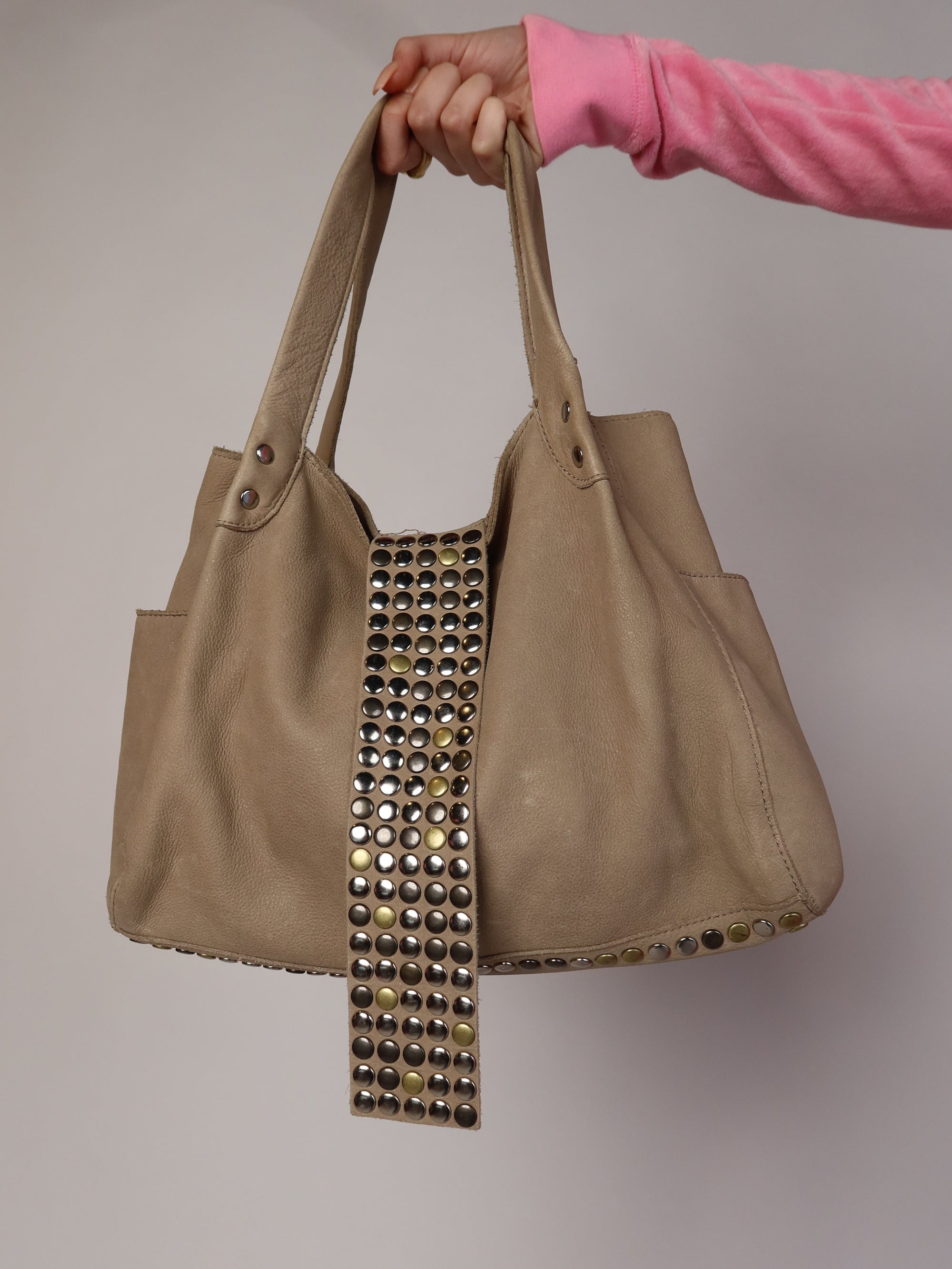 Authentic vintage Y2K slouchy tote shoulder bag made of real taupe leather, featuring a prominent wide front strap embellished with rows of metallic silver and gold studs, shown held up against a plain background.